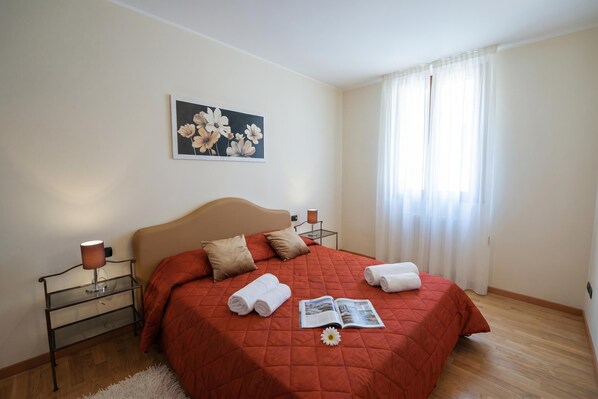 A neatly made bed with a red quilt, a bedside lamp, and a framed floral artwork on the wall.