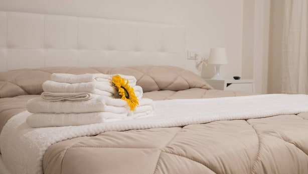 Ein ordentlich gemachtes Bett mit einer beigen Decke, einer weißen, flauschigen Wolldecke und einem Stapel weißer Handtücher mit einer gelben Blume.