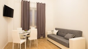 Ein kleiner, moderner Wohnraum mit einem grauen Sofa, weißen Stühlen, einem kleinen Tisch und einem Fernseher sowie einem Fenster mit Vorhängen.