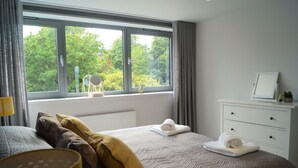 A hotel room with a large window, a bed with a patterned bedspread, two pillows, a bedside table with a lamp, and a dresser with a mirror and a small plant. - NEW! Luxury Apartment in Truro Centre With Cathedral Views! (Truro)