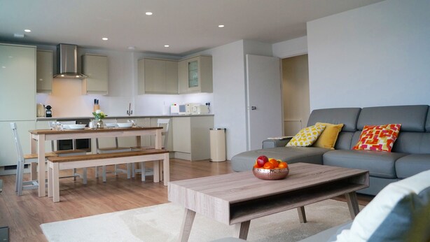 A modern living room with a grey sofa, a wooden coffee table, and a dining area with a bench and table. - NEW! Luxury Apartment in Truro Centre With Cathedral Views! (Truro)