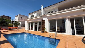 Une piscine avec une terrasse carrelée, un espace extérieur couvert aménagé en salon, et un bâtiment doté de grandes portes vitrées.