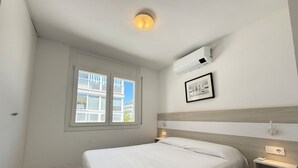 Une chambre à coucher comprenant un lit, une fenêtre, un appareil de climatisation, une table de chevet et une étagère fixée au mur.