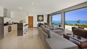 A modern kitchen with a central island, a dining area with bar stools, a large grey sofa, and a view of the ocean. - Tranquil Residence 3 (Koh Samui)