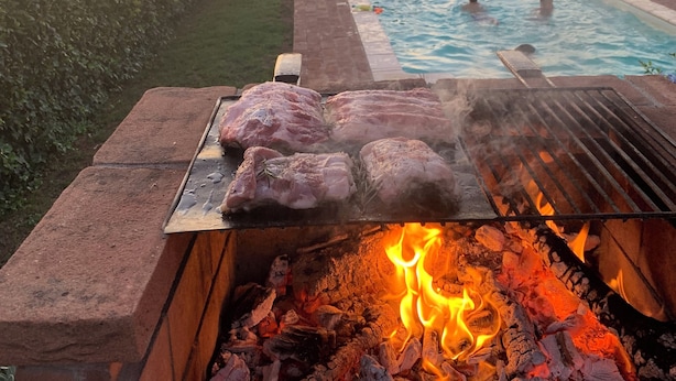 Uma churrasqueira com carne assando sobre lenha, uma piscina ao fundo e um céu ao pôr do sol.