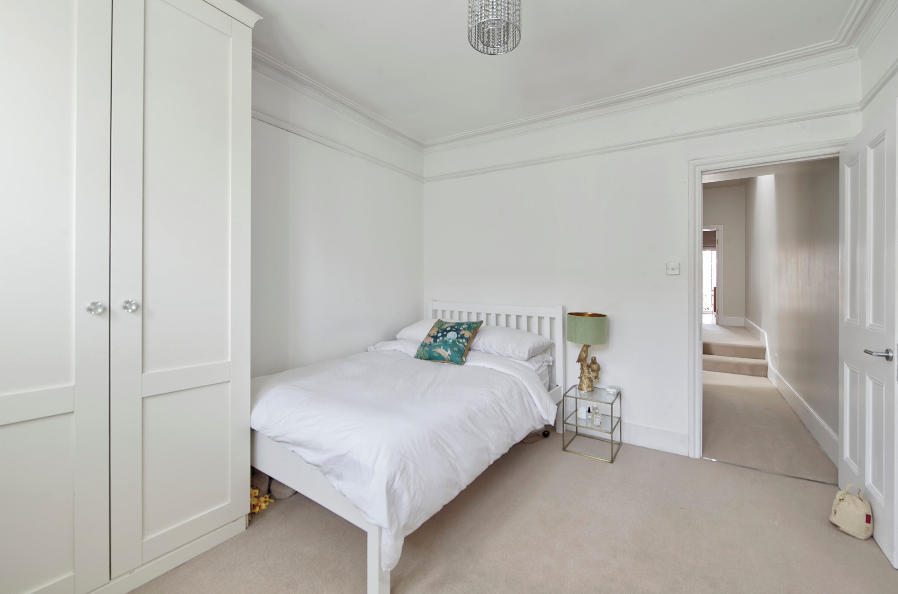 Un dormitorio con una cama blanca, una mesita de noche con una lámpara y una puerta que da a otra habitación.