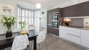 Een moderne keuken met een inbouwoven, een eetgedeelte met een zwarte tafel en witte stoelen, en een groot raam met zonwering.
