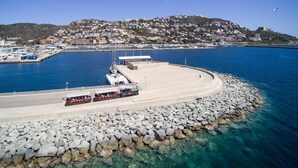 A coastal area with a marina, a rocky breakwater, and a hillside town in the background. - PUIG ROM 115 1 2  Roses (Rosas / Roses)