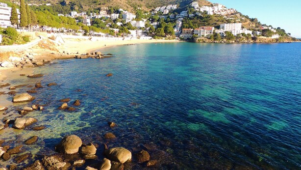 A coastal town with clear blue waters, rocky shoreline, and buildings on a hillside. - PUIG ROM 115 1 2  Roses (Rosas / Roses)