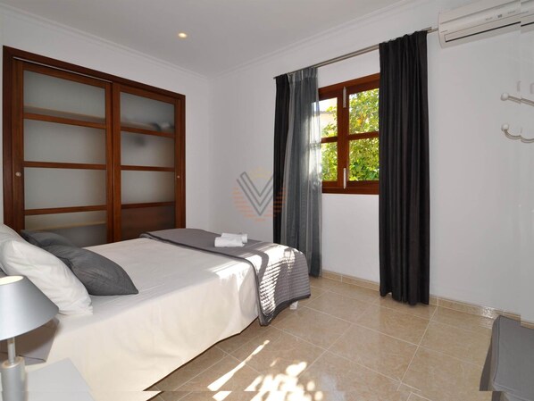 Una habitació d'hotel amb un llit, una taula de treball, una finestra amb cortines i un aparell d'aire condicionat.
