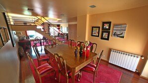 Uma sala de jantar com uma mesa de madeira, cadeiras vermelhas e um tapete vermelho. Uma escada ao fundo leva a outro cômodo.