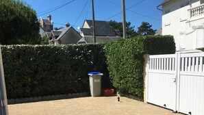 A paved outdoor area with a trash bin, a white gate, and a hedge.