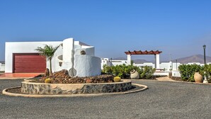 A modern building with a red garage door, a white wall with a circular cutout, and a garden with cacti and potted plants. - Villa the path of the majorero (Antigua)