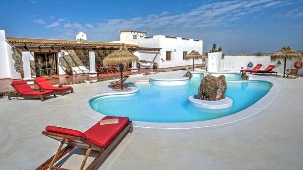 A pool area with red lounge chairs, a thatched umbrella, and a white building with a stone facade. - Villa the path of the majorero (Antigua)