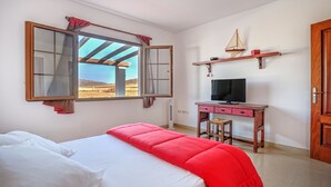A bedroom with a bed, a desk with a TV, a window with curtains, and a shelf with a sailboat model. - Villa the path of the majorero (Antigua)