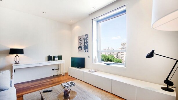 A modern living room with a large window, a flat-screen TV, a white bench, and a coffee table.