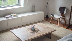 A room with a wooden coffee table, a white bench with books, a chair with a black and white cushion, and a floor lamp.