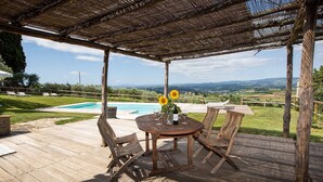 A wooden deck with a pergola, a round table with chairs, a pool, and a scenic view of hills and a distant town. - Lovely apartment for 6 people with A/C, pool, WIFI, TV and panoramic view, close to San Gimignano (Poggibonsi)