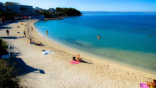 A beach with clear blue water, people sunbathing, and buildings in the background. - Split area studio apartment (Podstrana)