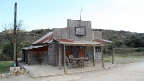 A rustic, single-story building with a corrugated metal roof and wooden walls, labeled as 'Shady Grove Station'. It features a small porch with wooden chairs and a table.