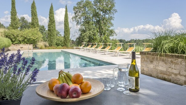 A poolside table with fruits, a bottle, and glasses, overlooking a pool and lounge chairs.