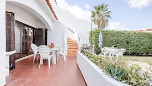 A modern outdoor patio with white chairs and a table, a staircase leading to an upper level, and a well-maintained hedge.