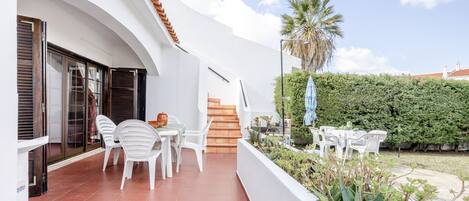 A modern outdoor patio with white chairs and a table, a staircase leading to an upper level, and a well-maintained hedge.