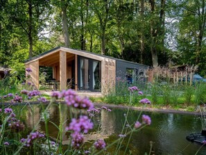 Eine moderne Holz-Blockhütte mit Glastür, umgeben von üppiger Vegetation und einem Teich.