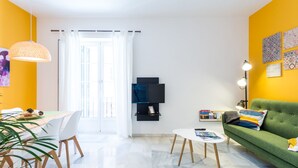 Un salon moderne avec un canapé vert, une table à manger avec des chaises, un téléviseur fixé au mur et une grande fenêtre avec des rideaux.