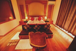 A hotel room with a wooden floor, a bed with heart-shaped decorations, a wooden chair, and a nightstand with lamps.