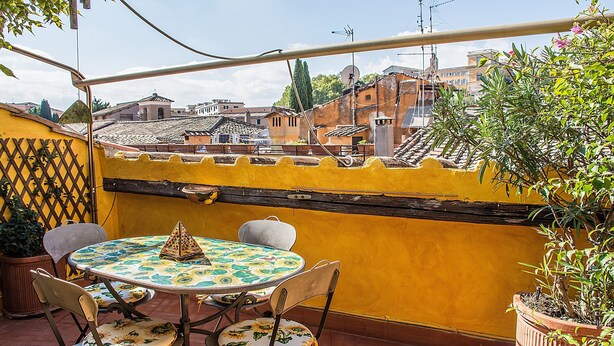 A patio with a round table and chairs, a yellow wall, and a view of buildings in the background. - Attic of great charm with terraces and panoramic views (Roma)