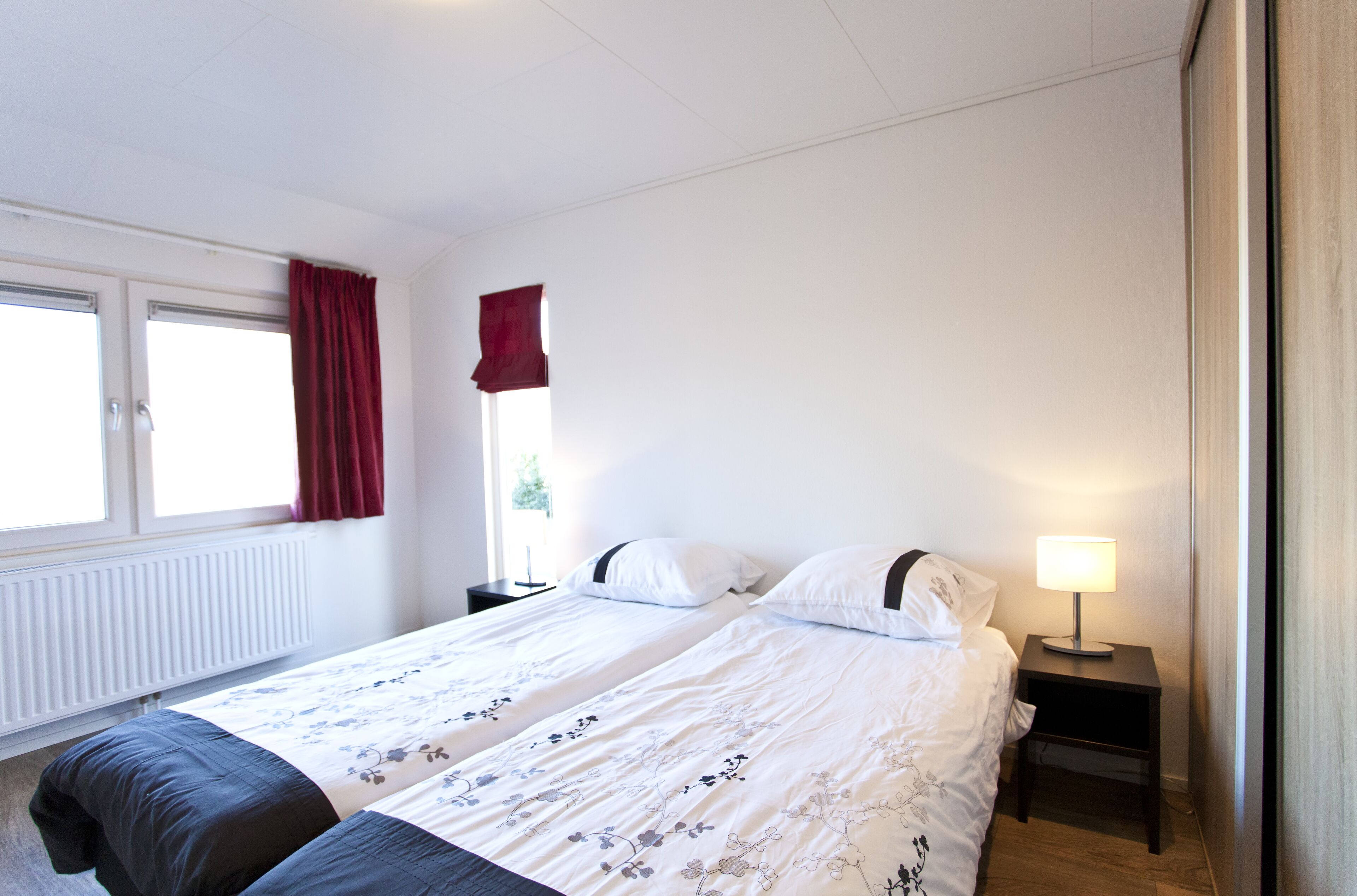 A hotel room with two beds, a nightstand with a lamp, a window with red curtains, and a radiator.