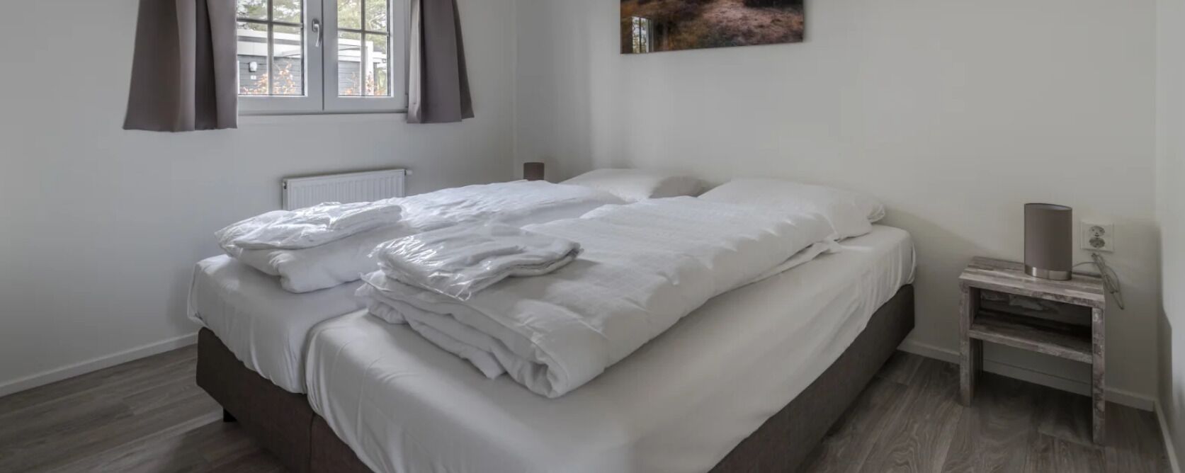 A neatly made bed with white linens, a bedside table with a lamp, and a window with curtains.