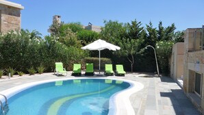 Une piscine à bordures courbes, entourée d’une terrasse en pierre. On y trouve quatre chaises longues vertes et un parasol blanc.