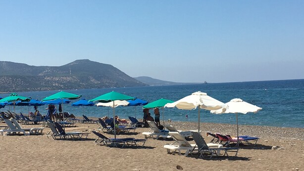 Une plage bordée de rangées de chaises longues et de parasols, un ciel dégagé et une chaîne de montagnes à l’horizon.