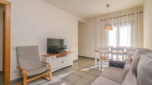 A living room with a sofa, a TV on a wooden cabinet, a dining table with chairs, and a window with curtains.