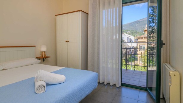 A hotel room with a bed, a nightstand with a lamp, a wardrobe, and a balcony with a view of buildings and greenery.