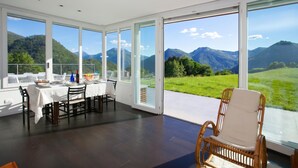 A dining area with a table set for two, large windows offering a mountain view, and a wicker chair. - Villa Intelvi, Cerano d'Intelvi, Italy (Cerano d'Intelvi)