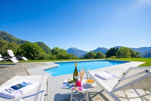 Escena junto a la piscina con sillas de descanso, una mesa con una botella, vasos y fruta, y montañas al fondo.