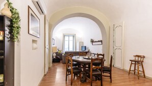 Ein Essbereich mit einem runden Holztisch und Stühlen, ein rundbogiges Durchgang zur nächsten Räumlichkeit und eine Pflanze an der linken Wand.