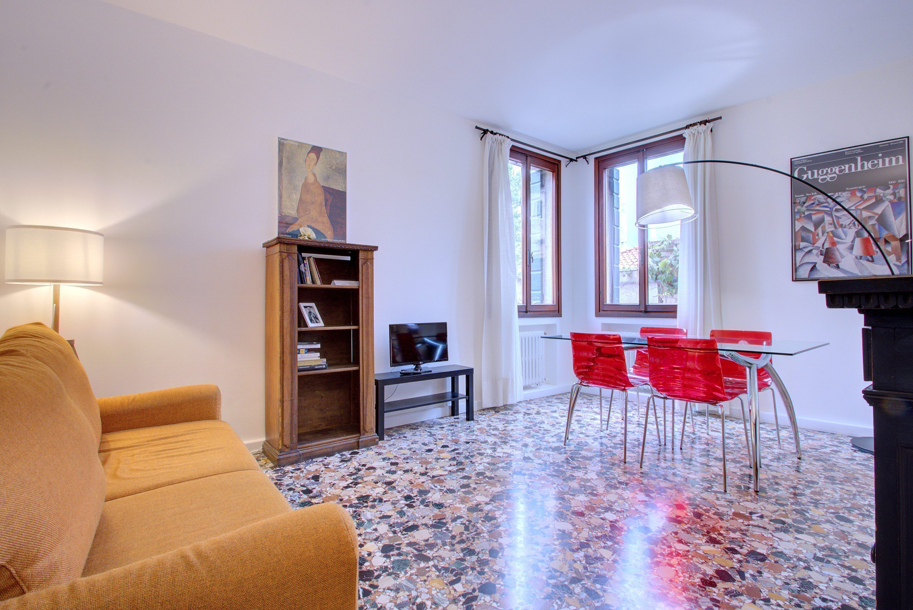 Un salon avec un canapé, un téléviseur, une bibliothèque, une table à manger avec des chaises rouges et une affiche accrochée au mur.