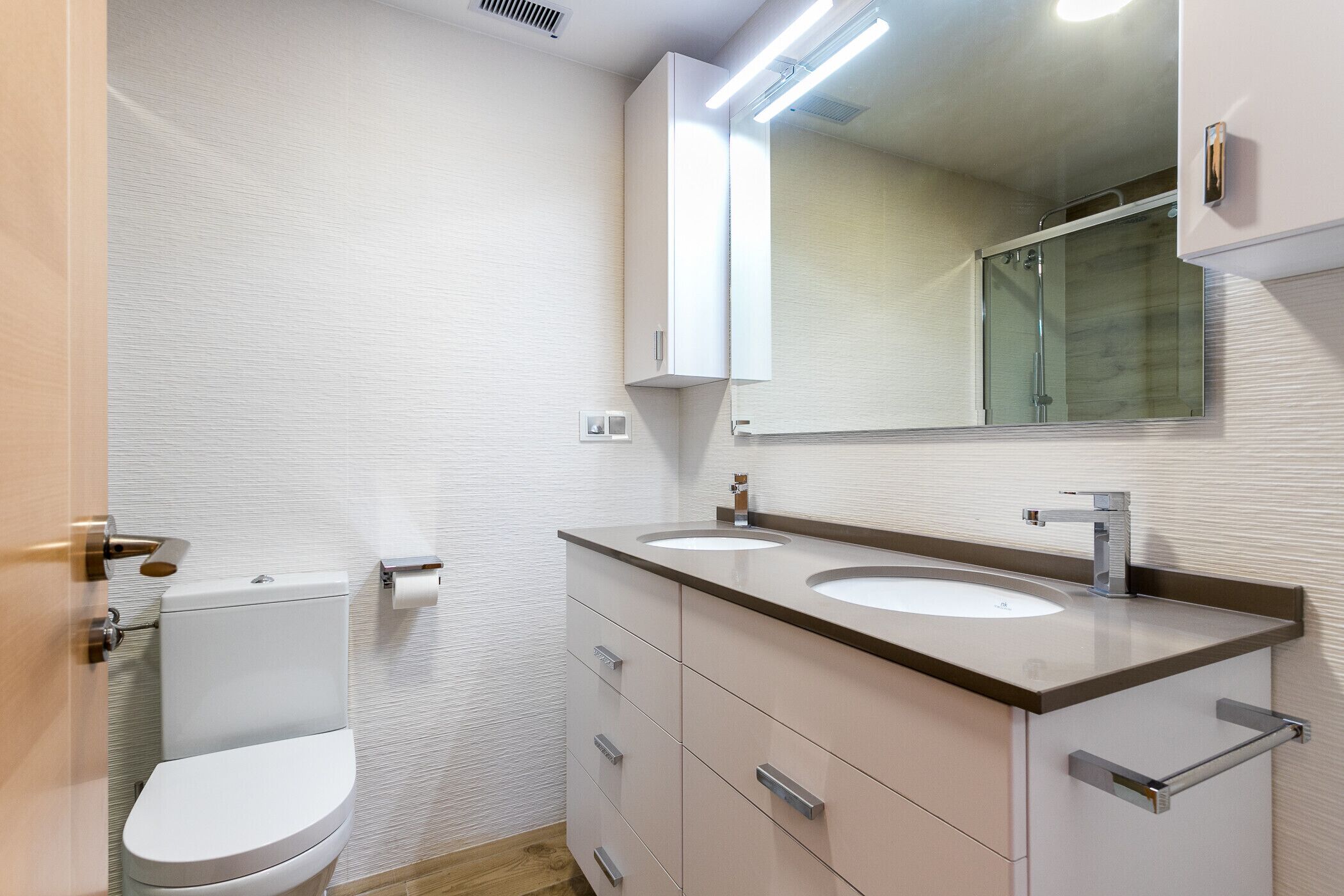 Moderni kylpyhuone, jossa on wc, kaksi pesuallasta, peili ja pyyhekoukku.