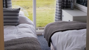 A bedroom with two beds, a bedside table with a lamp, a window with curtains, and a view of a grassy area.