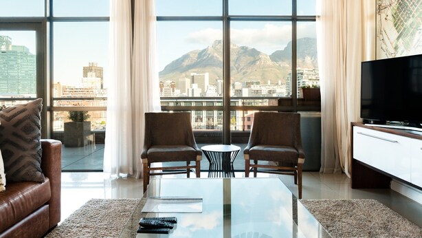 A modern living room with a large window, two armchairs, a coffee table, and a flat-screen TV. - Hillhouse Penthouse 7th Floor (Cape Town)