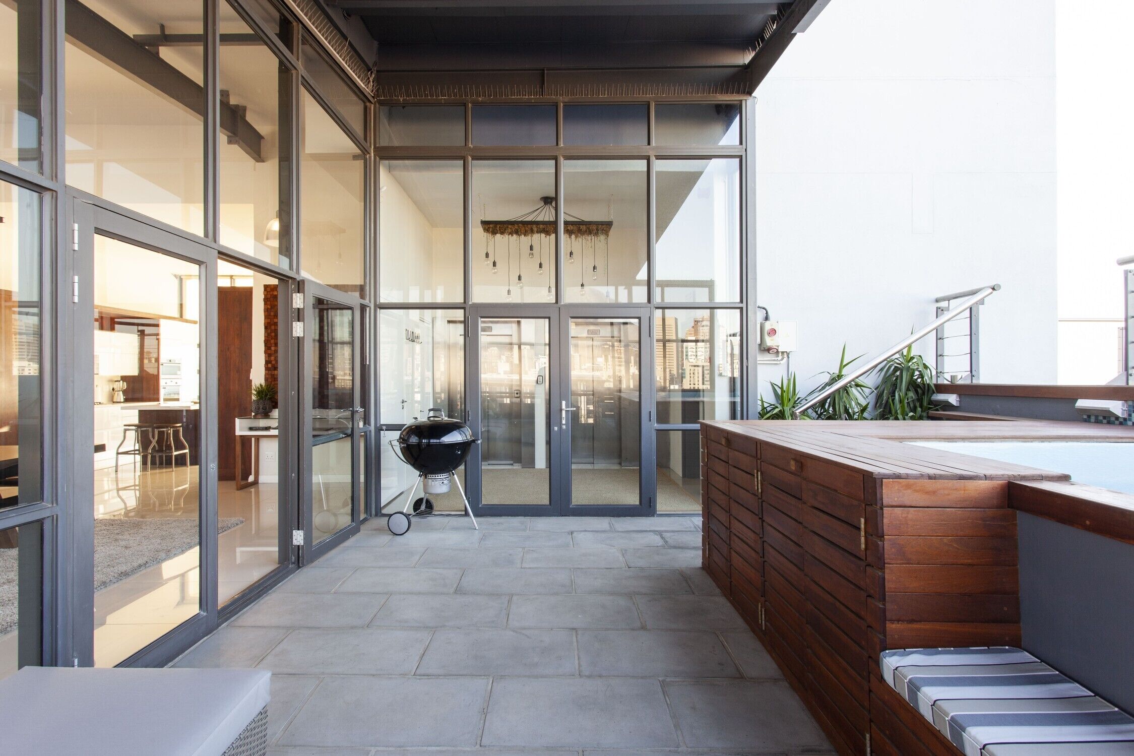A modern outdoor patio area with a barbecue grill, a wooden bench, and a glass door leading to an interior space.