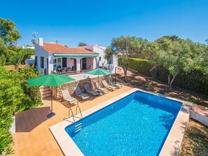 A swimming pool with lounge chairs and umbrellas, a stone wall, and a white building with a red roof.