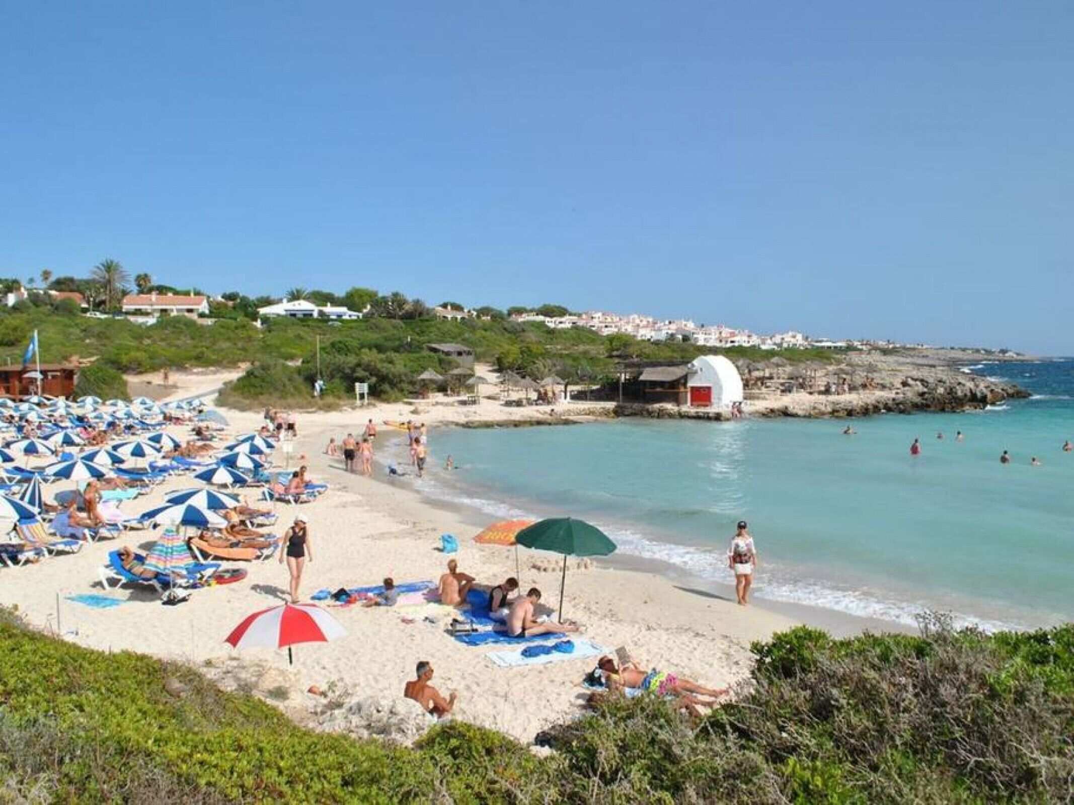 Ein Strand mit vielen Liegestühlen und Sonnenschirmen, ein kleines weißes Gebäude und Menschen, die im Wasser schwimmen.