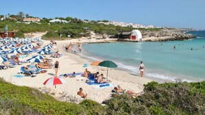 Ein Strand mit vielen Liegestühlen und Sonnenschirmen, ein kleines weißes Gebäude und Menschen, die im Wasser schwimmen.