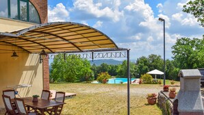 Una veranda coperta con tavolo e sedie, un barbecue e vista su una piscina e montagne sullo sfondo.