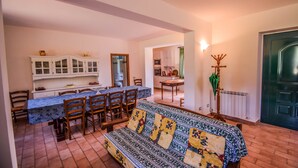 Una zona pranzo con un tavolo lungo e sedie, un divano con cuscini decorati, e una cucina sullo sfondo.